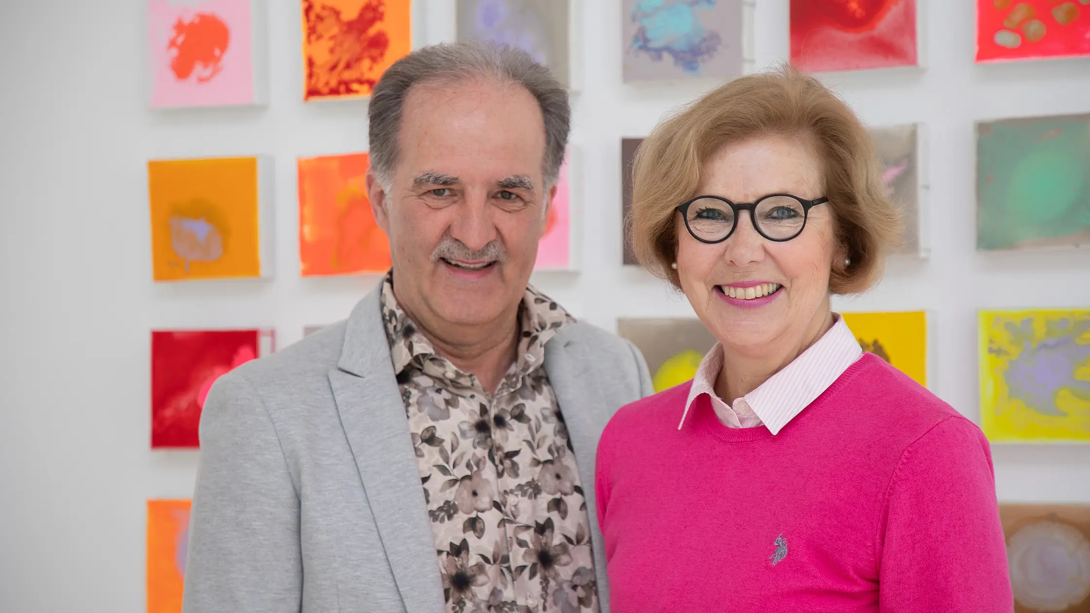Zwei Patientenfürsprecher:innen stehen nebeneinander vor einer Wand mit farbigen, quadratischen Kunstwerken und blicken zur Kamera.