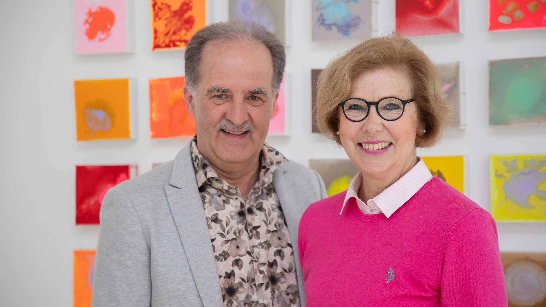 Zwei Patientenfürsprecher:innen stehen nebeneinander vor einer Wand mit farbigen, quadratischen Kunstwerken und blicken zur Kamera.
