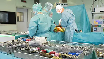 Mehrere Personen in grüner OP-Kleidung stehen um einen Operationstisch und führen gemeinsam eine Hüftoperation durch. Im Vordergrund sind Instrumententische mit chirurgischem Material zu sehen.