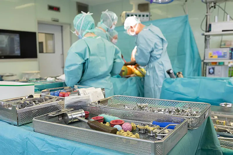 Mehrere Personen in grüner OP-Kleidung stehen um einen Operationstisch und führen einen chirurgischen Eingriff durch. Im Vordergrund sind Instrumententische mit medizinischem Material zu sehen.
