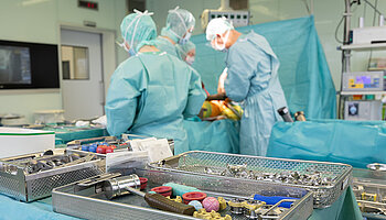 Mehrere Personen in grüner OP-Kleidung stehen um einen Operationstisch und führen gemeinsam eine Hüftoperation durch. Im Vordergrund sind Instrumententische mit chirurgischem Material zu sehen.