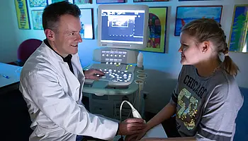 Arzt führt eine Ultraschalluntersuchung am Unterarm eines Mädchens durch; das Ultraschallgerät steht daneben.