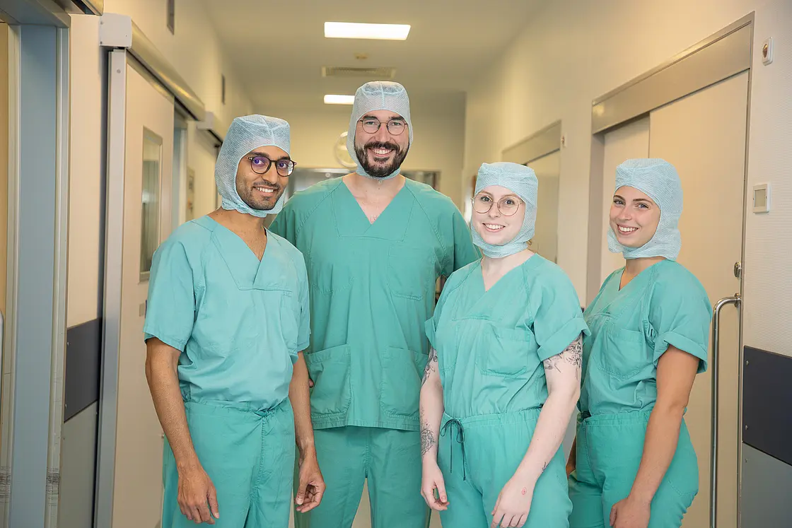 Vier Personen in grüner OP-Kleidung und Hauben stehen in einem Krankenhausflur und schauen in die Kamera. Sie tragen sterile Arbeitskleidung und lächeln.