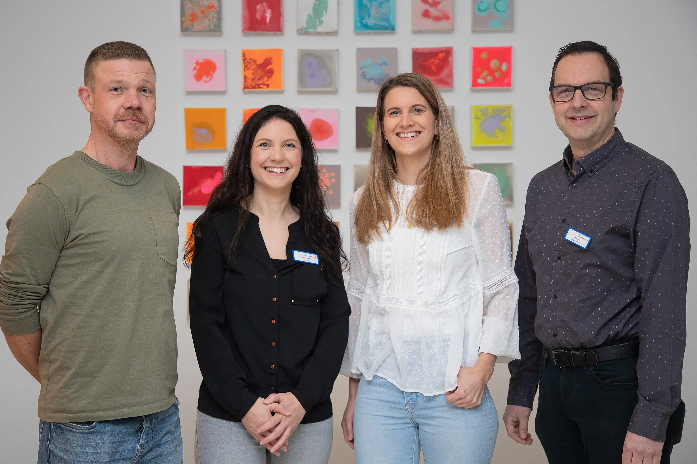 Vier Mitarbeitende des Hygieneteams stehen nebeneinander in einem Innenraum und blicken in die Kamera; im Hintergrund sind farbige Wandbilder zu sehen.