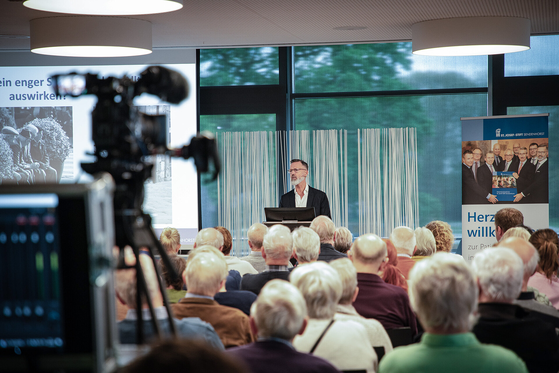 Referent (Dr. Christian Brinkmann) hält einen Vortrag vor Publikum in einem Veranstaltungsraum, im Vordergrund ist eine Kamera zu sehen.