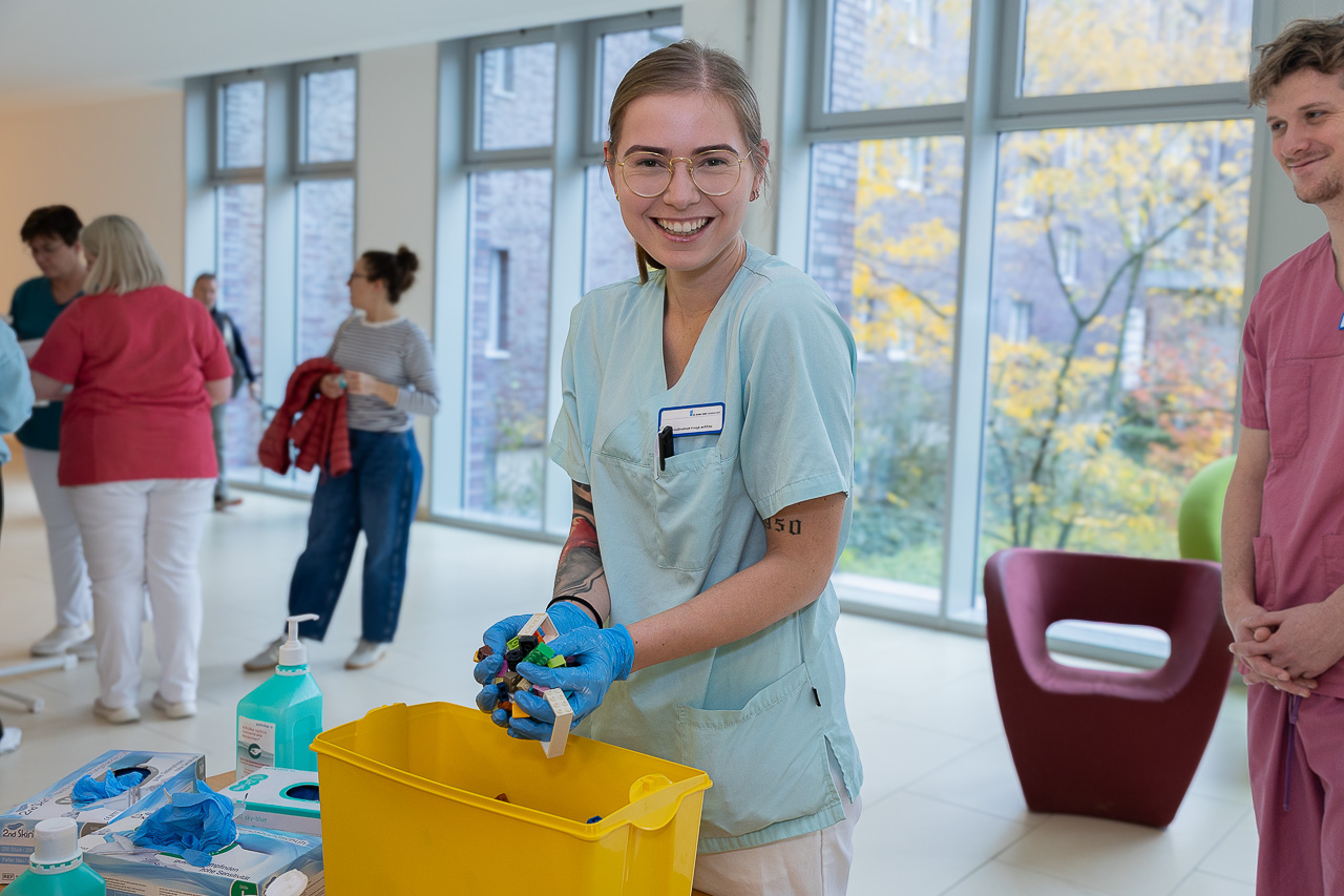 Eine Mitarbeiterin in Arbeitskleidung und Handschuhen zeigt im Rahmen der Aktion Saubere Hände gesammelte Handschuhe an einem Informationsstand; im Hintergrund stehen weitere Personen im Klinikflur.