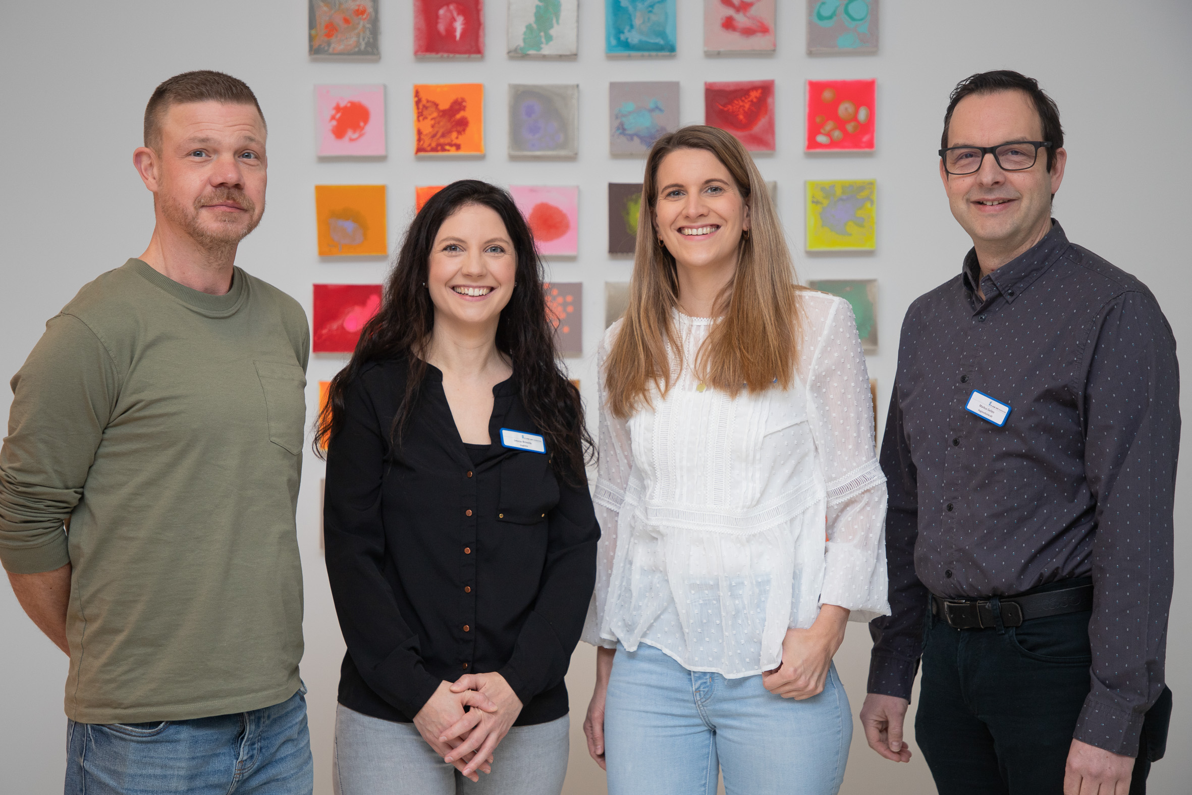 Vier Mitarbeitende des Hygieneteams stehen nebeneinander in einem Innenraum und blicken in die Kamera; im Hintergrund sind farbige Wandbilder zu sehen.