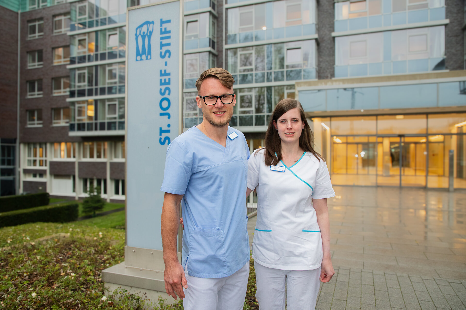 Ein Mann und eine Frau stehen vor einem warm beleuchteten Krankenhauseingang, hinter ihnen ein Krankenhausschild.