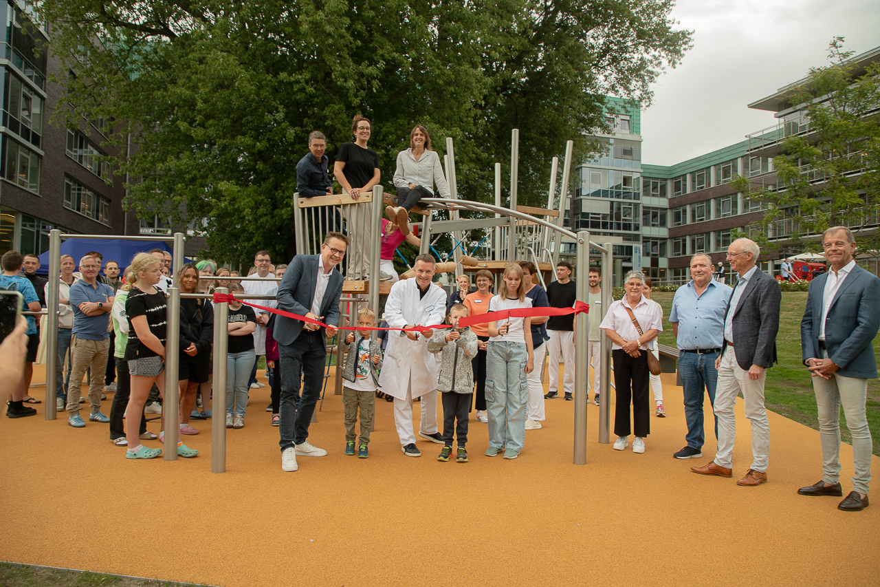 Viele ältere und junge Menschen stehen auf einem neuen Spielplatz, in der Mitte ein Arzt im weißen Kittel, der symbolisch ein rotes Band mit einer Schere durchschneidet.