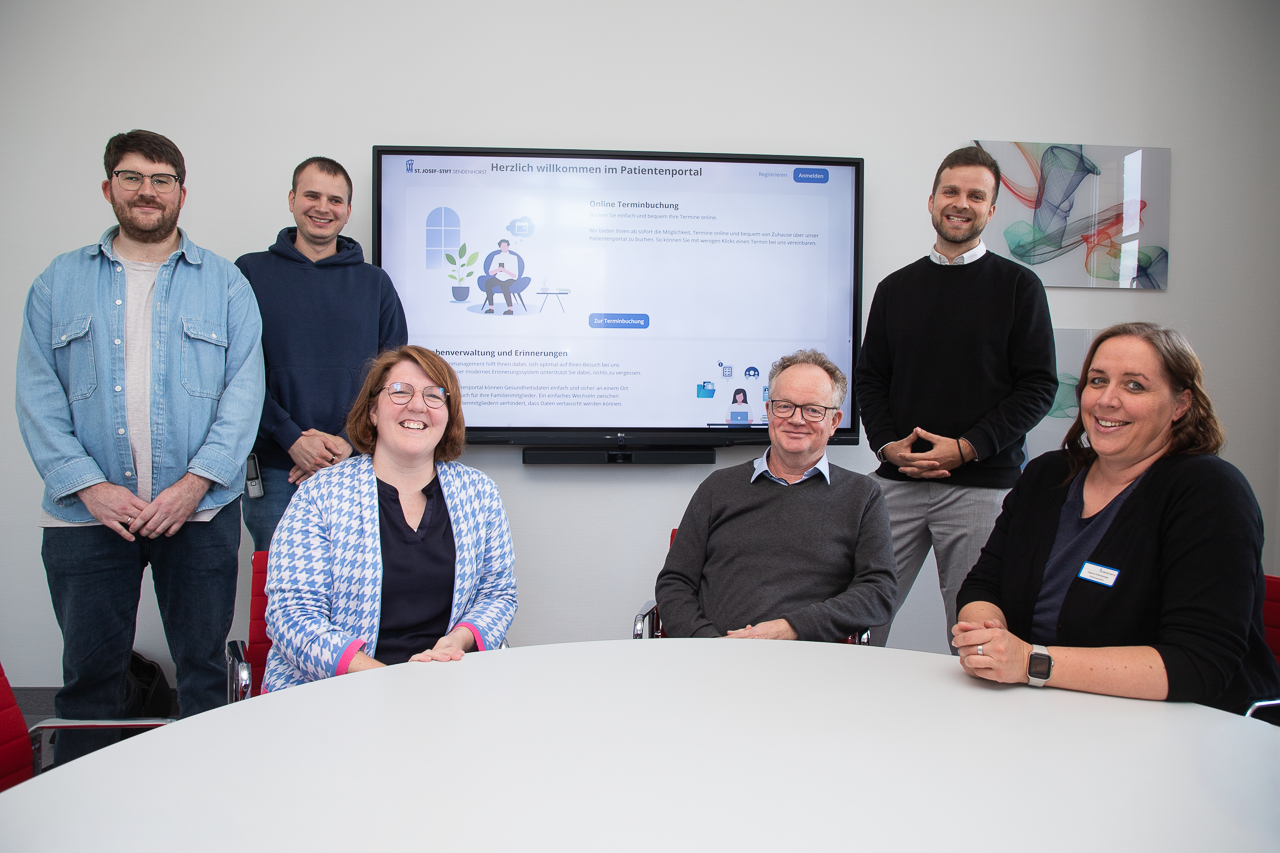 Mitarbeitende sitze am Tisch vor einem Bildschirm, auf dem das Patientenportal zu sehen ist.