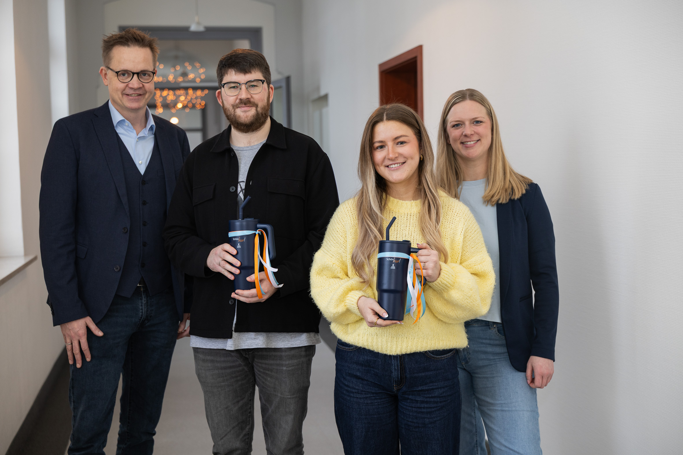 Vier Menschen stehen in einem Flur nebeneinander und lächeln, die beiden Auszubildenden in der Mitte halten jeweils einen blauen Trinkbecher in der Hand.