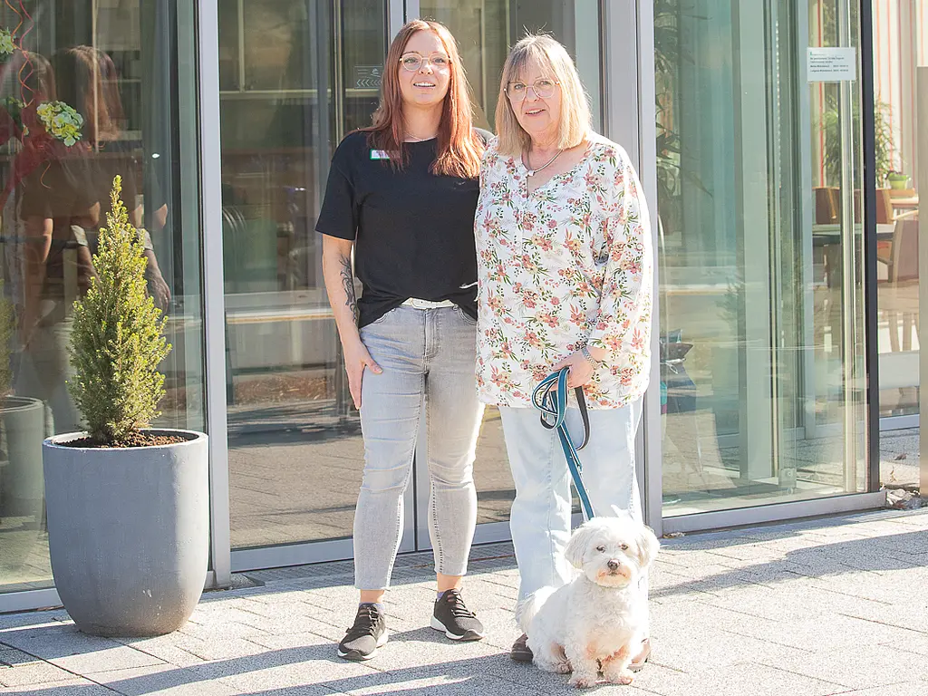 Eine jüngere und eine ältere Frau, Tochter und Mutter, stehen mit einem kleinen Hund an der Leine vor dem Eingang der Pflegeeinrichtung in Albersloh.