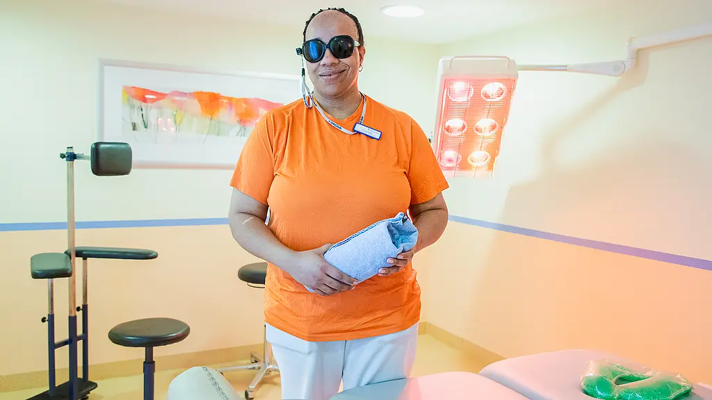 Eine Frau steht mit einem gerollten Handtuch in der Hand vor einer Massageliege, hinter ihr sind eine leuchtende Wärmelampe und ein Massagestuhl zu sehen.