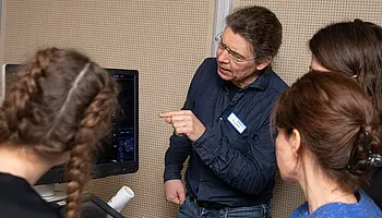 Ein Arzt erklärt etwas und zeigt mit einem Finge auf einen Bildschirm, auf dem Ultraschallbilder zu sehen sind.