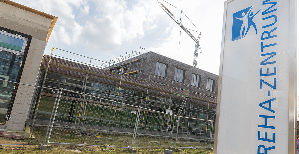Außenansicht des Reha-Neubaus mit Baugerüst und Kran; im Vordergrund ein Schild mit der Aufschrift „Reha-Zentrum“, das Gelände ist noch abgesperrt