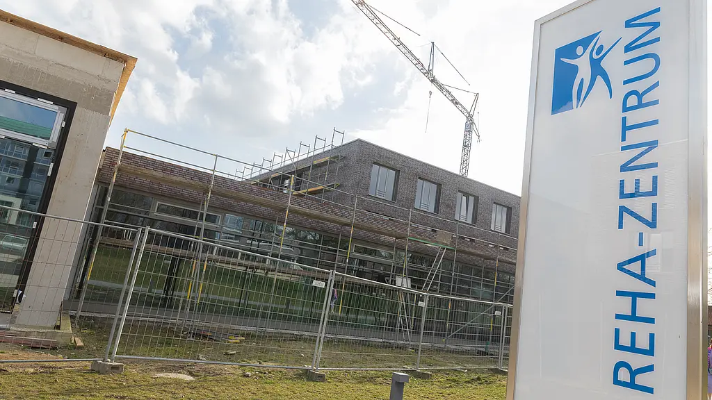 Außenansicht des Reha-Neubaus mit Baugerüst und Kran; im Vordergrund ein Schild mit der Aufschrift „Reha-Zentrum“, das Gelände ist noch abgesperrt