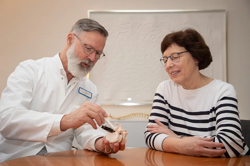 Ein Arzt erklärt einer Patientin eine Wirbelsäulenoperation am Modell, dafür zeigt er mit einem Stift auf das Modell eines Wirbelsäulenstücks in seiner Hand.
