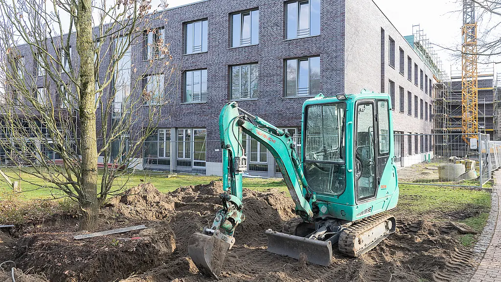 Bagger arbeitet im Außenbereich vor dem Neubau des Reha-Zentrums; daneben ein mehrgeschossiges Gebäude mit Backsteinfassade und eingerüsteten Bereichen im Hintergrund