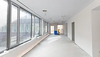 Heller, langer Flur im Neubau der Reha mit großen Fensterfronten links, noch unfertiger Decke mit sichtbaren Kabelanschlüssen und leerem Boden; Baumaterialien stehen im Hintergrund